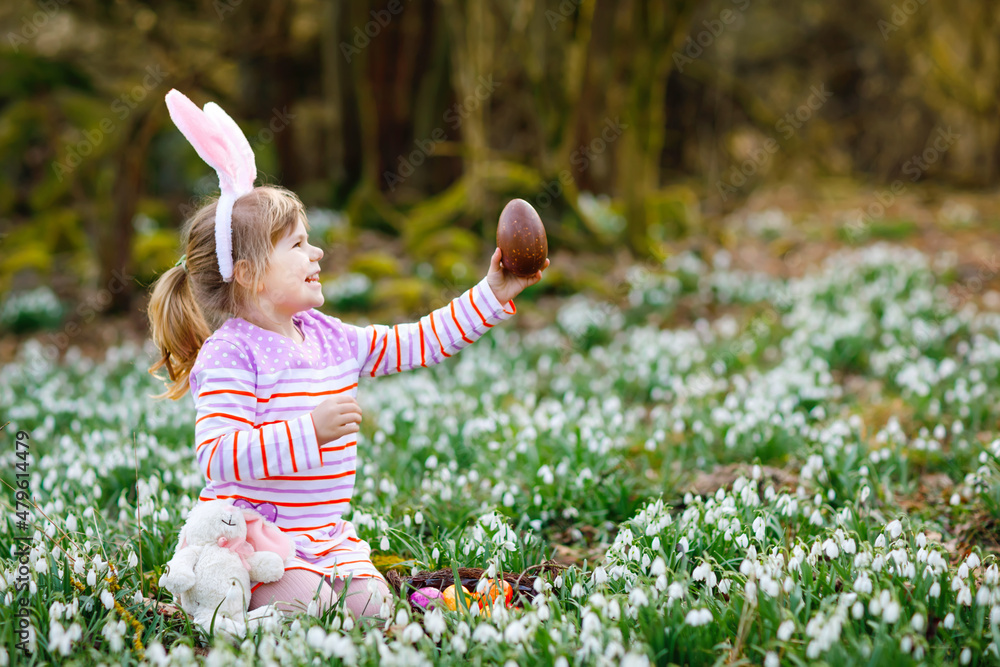 Little girl with Easter bunny ears making egg hunt in spring forest on sunny day, outdoors. Cute happy child with lots of snowdrop flowers, huge chocolate egg and colored eggs. Little girl with Easter bunny ears making egg hunt in spring forest on sunny day, outdoors. Cute happy child with lots of snowdrop flowers, huge chocolate egg and colored eggs.