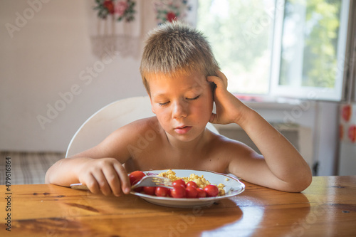 Blond child in domestic kitchen eat healthy food, organic vegan fritatta  with vegetables. Portrait of young boy looking fed up with plate of healthy food photo