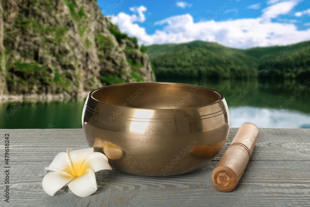 Golden singing bowl, mallet and flower on grey wooden table against mountain landscape