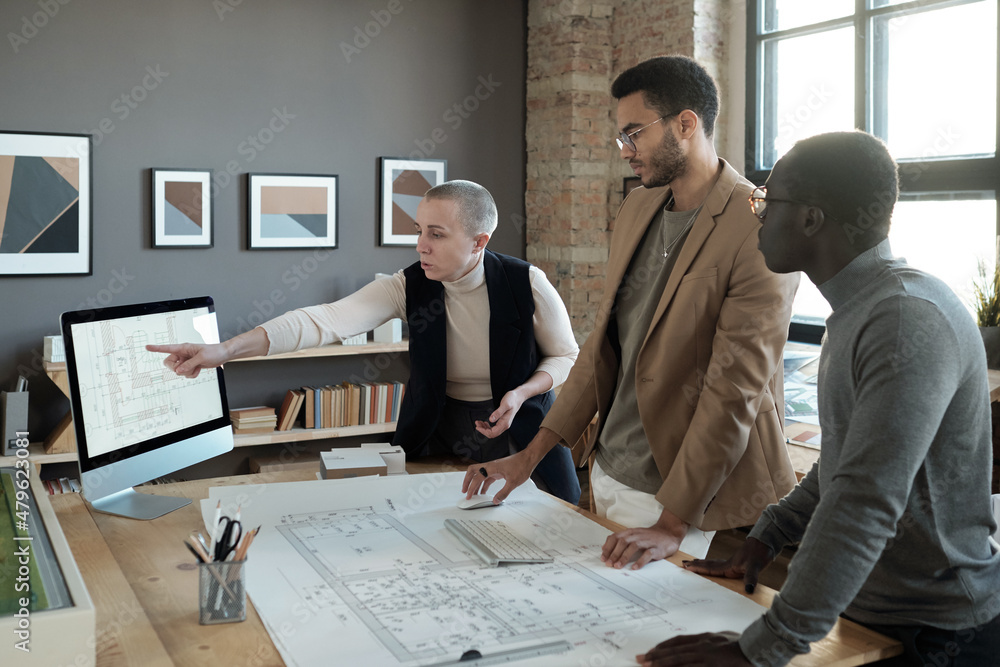 Mature businesswoman pointing at sketch on computer screen while explaining it to intercultural male colleagues Mature businesswoman pointing at sketch on computer screen while explaining it to intercultural male colleagues