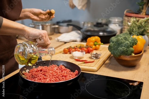 Hands of woman pouring olive oil into frying pan with raw minced meat while cooking traditional italian pasta at home