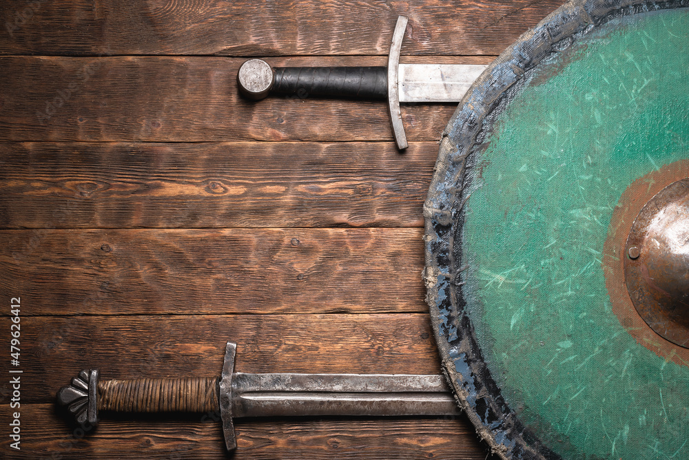 Fototapeta premium Two swords and the shield on the old medieval table flat lay concept background with copy space.