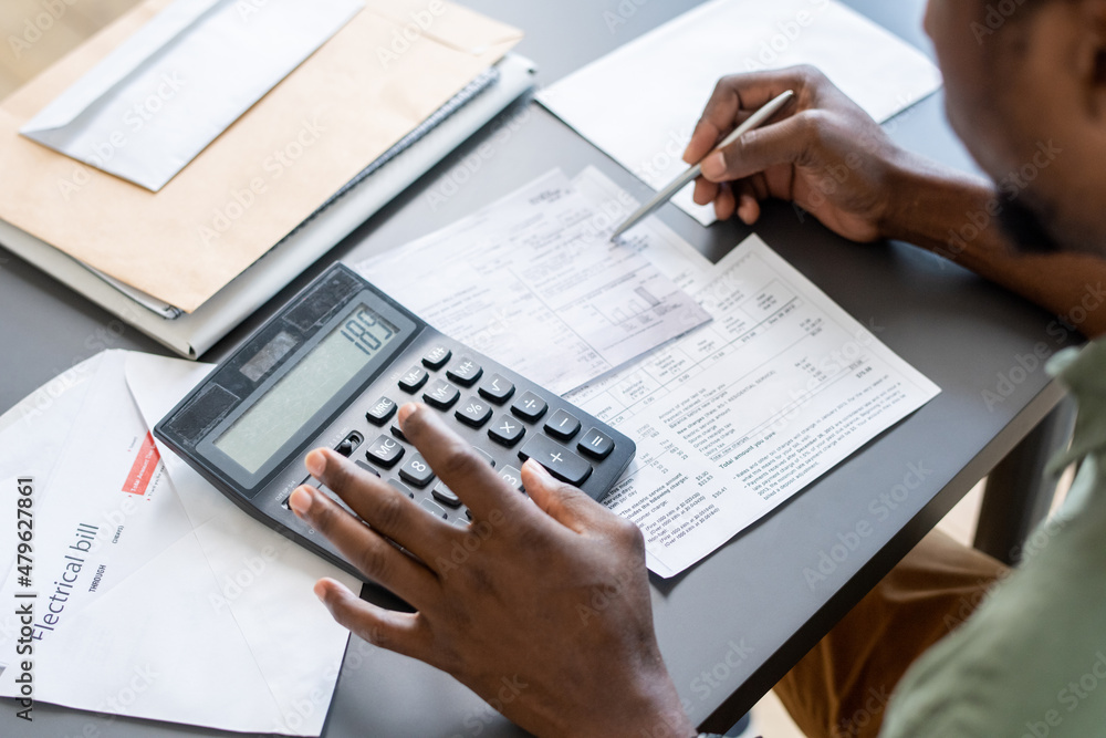 Hands of African man pressing buttons of calculator while checking data in financial papers and unpaid bills by table - obrazy, fototapety, plakaty 