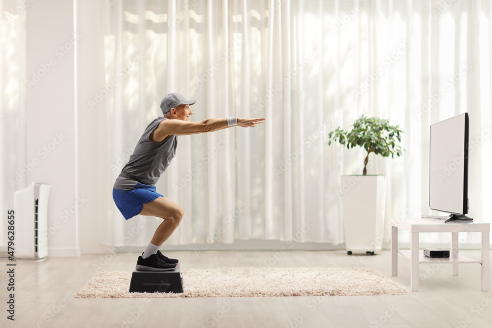 Elderly man exercising squats on a step aerobic platform in front of tv