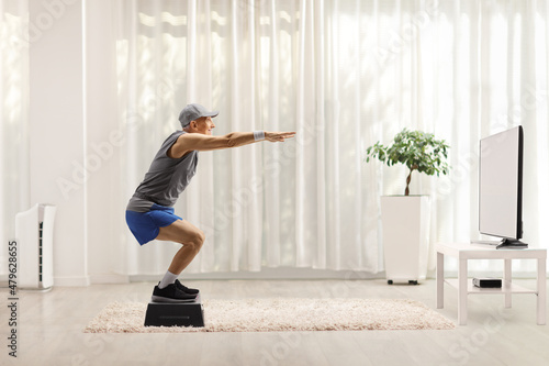 Fototapeta Naklejka Na Ścianę i Meble -  Elderly man exercising squats on a step aerobic platform in front of tv