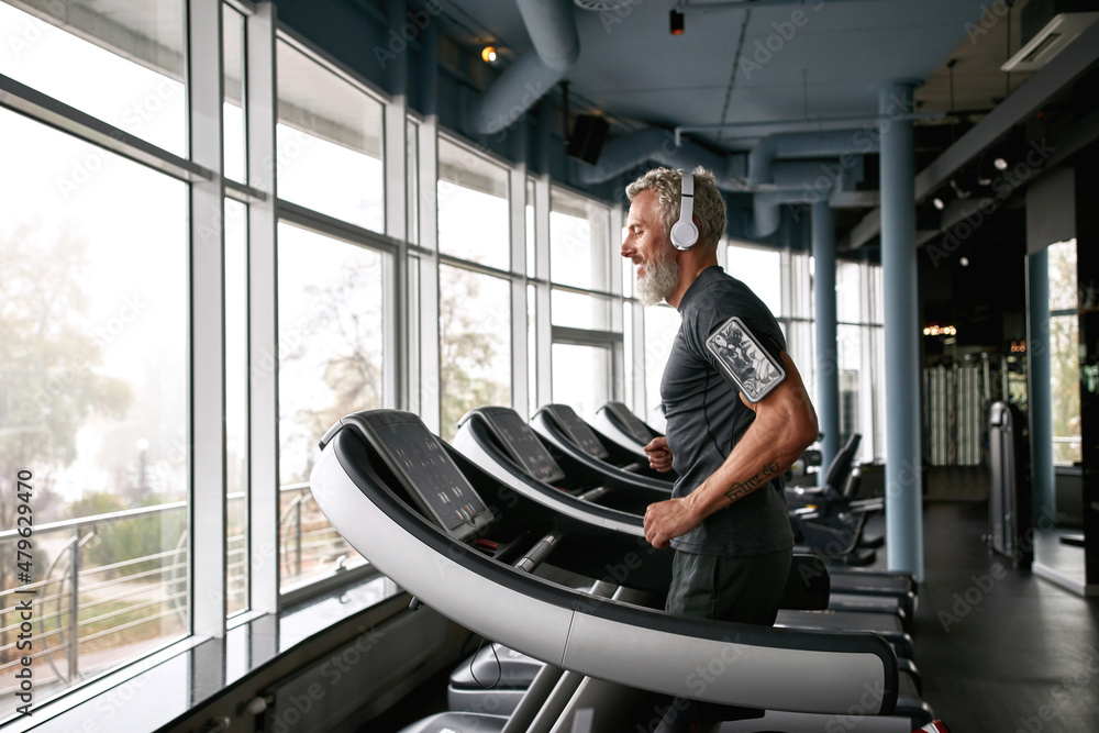 Fototapeta premium Senior man in gray t-shirt and white headphones in gym