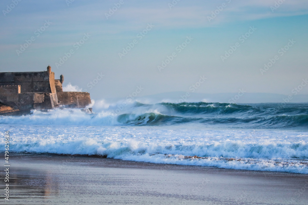 Large waves crash into a fortress near a beach. Carcavelos beach in Portugal