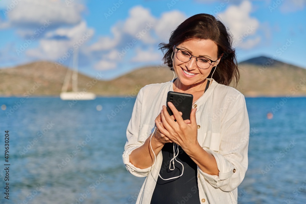 Middle-aged woman relaxing on beach wearing headphones with smartphone