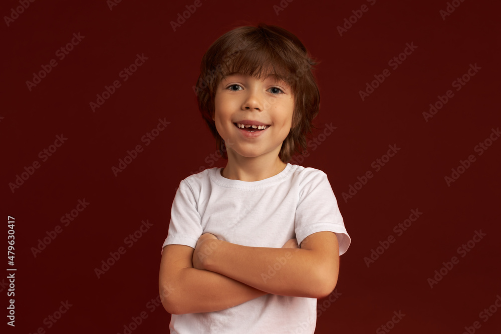 Cute lovely hairy-headed kid in white t-shirt standing with hands folded, looking at camera with charming toothy smile showing his new permanent upper teeth, posing against maroon wall Cute lovely hairy-headed kid in white t-shirt standing with hands folded, looking at camera with charming toothy smile showing his new permanent upper teeth, posing against maroon wall