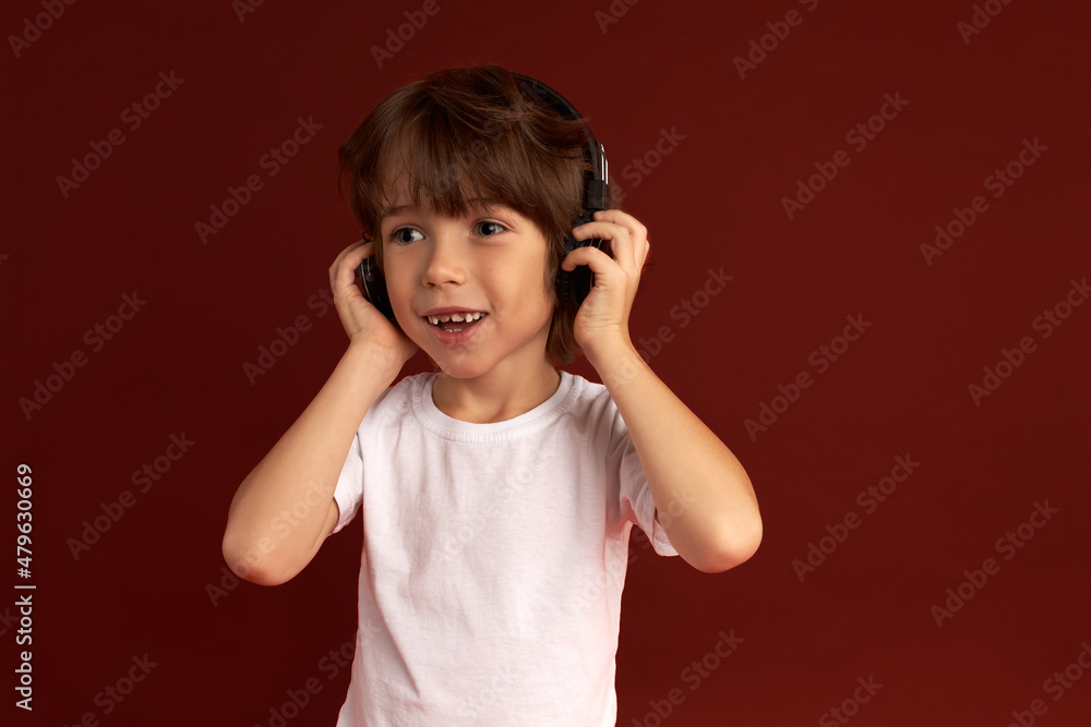 Kids and technology. Adorable little boy standing against red wall in wireless stereo headphones listening to Mozart, having excited and happy face expression, putting hands on them Kids and technology. Adorable little boy standing against red wall in wireless stereo headphones listening to Mozart, having excited and happy face expression, putting hands on them