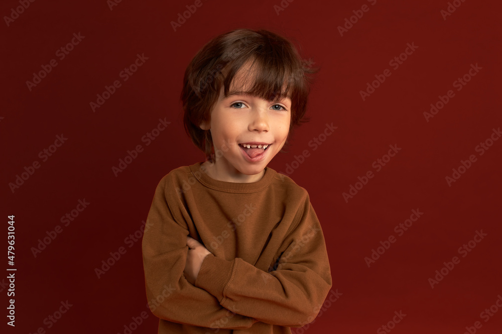 Adorable good-looking little naughty boy sticking tongue, teasing you looking at camera with his beautiful blue eyes, keeping arms crossed, wearing brown sweater, isolated on red studio wall Adorable good-looking little naughty boy sticking tongue, teasing you looking at camera with his beautiful blue eyes, keeping arms crossed, wearing brown sweater, isolated on red studio wall