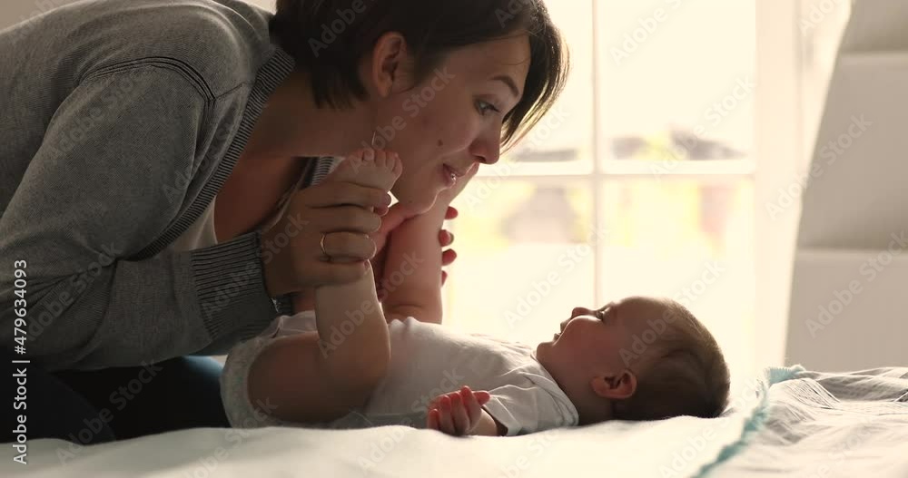 Loving mom kisses tiny toes of her cute 0-6 baby girl, close up. Smiling infant lying on bed enjoy mum caress and playtime looks satisfied. Babyhood, happy motherhood, mothers sincere love concept Loving mom kisses tiny toes of her cute 0-6 baby girl, close up. Smiling infant lying on bed enjoy mum caress and playtime looks satisfied. Babyhood, happy motherhood, mothers sincere love concept