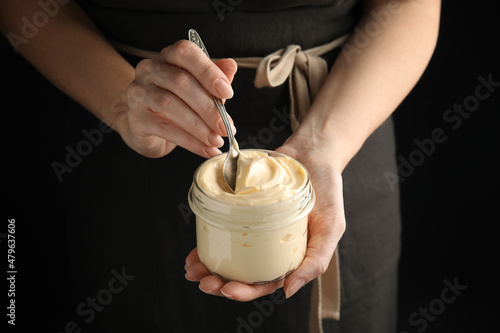 Woman holding spoon and jar of delicious mayonnaise on black background, closeup photo