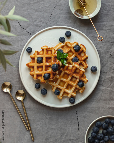Waffles with addition of fresh blueberries and honey on a white plate on table with natural grey linen tablecloth close up view action shot girl pouring honey on waffles
