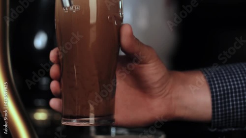 Barman in plaid shirt pours a pint of cold dark beer into a clear glass, the foam slowly settles. High quality 4k footage