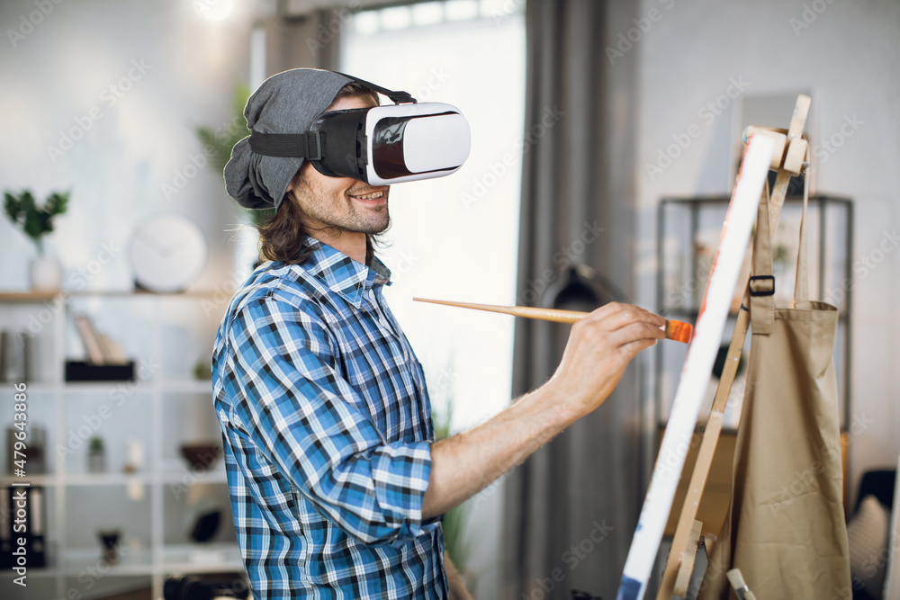 Caucasian male painter wearing VR headset while painting with brush on canvas at studio. Young man using innovative technology for creative process indoors.