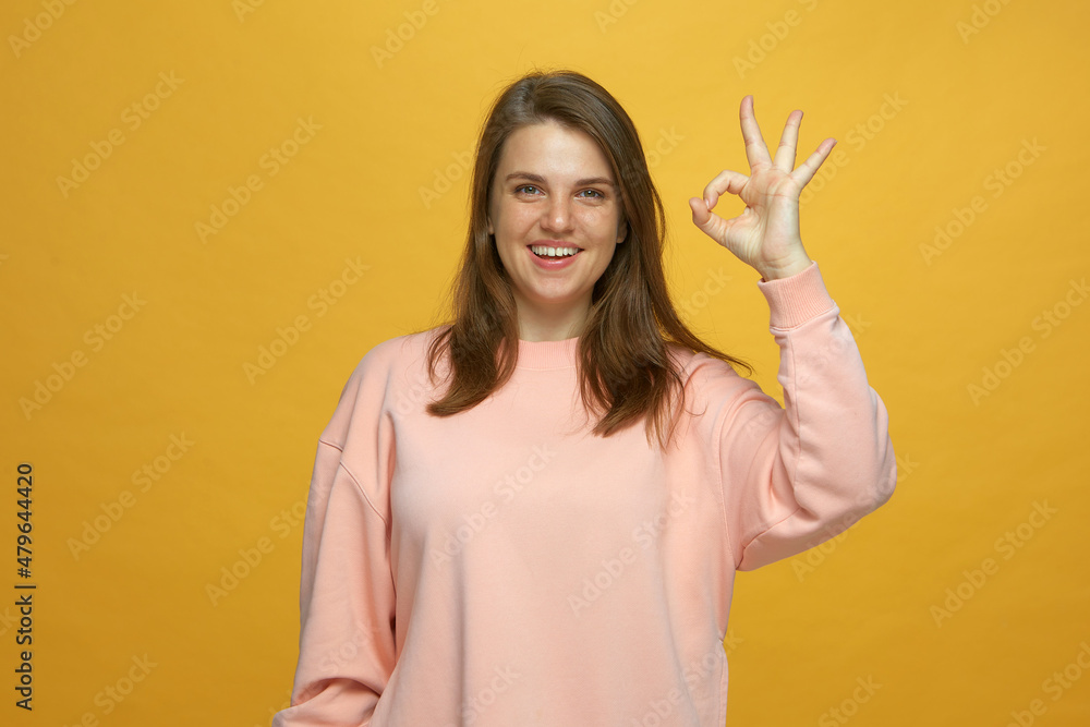 Happy young girl gesturing ok smiling looking at camera showing okay sign satisfied with service on yellow background Happy young girl gesturing ok smiling looking at camera showing okay sign satisfied with service on yellow background