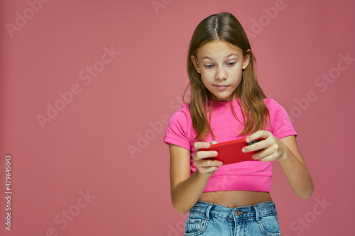 Puzzled child girl online gaming, playi using mobile phone app on pink background. Gadgets addiction in childhood