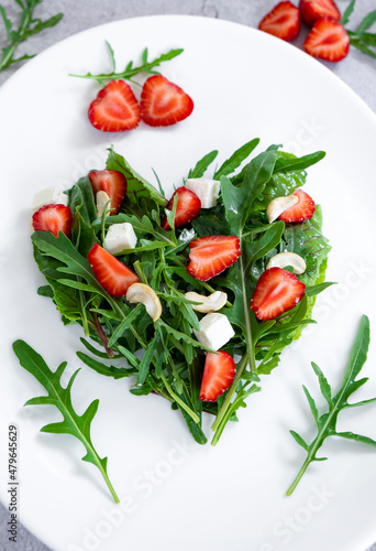 Delicious salad of strawberry with mix of herbs, arugula, spinach leaves, feta cheese and cashew nuts in heart shape on white plate. Vegetarian food and raw food diet.