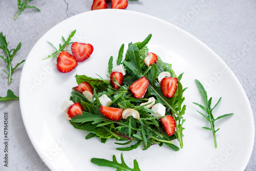 Delicious salad of strawberry with mix of herbs, arugula, spinach leaves, feta cheese and cashew nuts in heart shape on white plate. Vegetarian food and raw food diet.