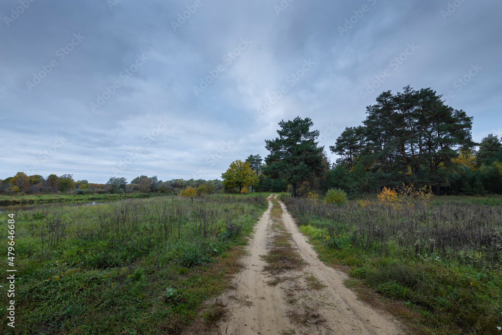 Dirt road along the river bank. Cloudy autumn weather. Rural landscape with road, river and autumn forest.