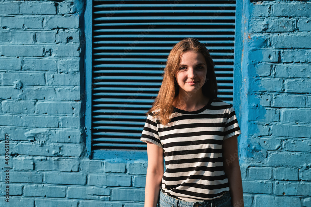 Portrait of a young lovely woman with blue eyes and freckles in a striped T-shirt and jeans against blue brick wall background. Portrait of a young lovely woman with blue eyes and freckles in a striped T-shirt and jeans against blue brick wall background.