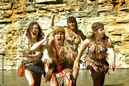 A group of woman dress up as a Neanderthal warriors. Their bodies and faces are covered with mud and dirt. Their facial expression is filled with anger as they wade through water