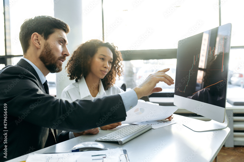 Naklejka premium Successful Caucasian man and African American woman, crypto traders, sitting in an office, using a computer, looking at charts, analyzing the crypto market, planning a strategy for good profit