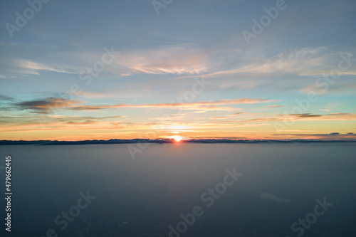 Aerial view of colorful sunset over white dense foggy clouds cover with distant dark silhouettes of mountain hills on horizon