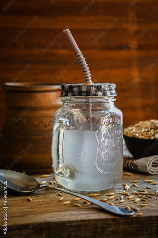 On the wooden kitchen table are a ceramic bowl of oat grains, a glass cup with oat kvass on a kitchen napkin, and an earthen jug. Village life. On the wooden kitchen table are a ceramic bowl of oat grains, a glass cup with oat kvass on a kitchen napkin, and an earthen jug. Village life.