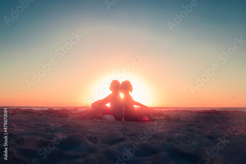 Pareja de enamorados sentados de espaldas en la playa a contraluz al amanecer Fotobehang