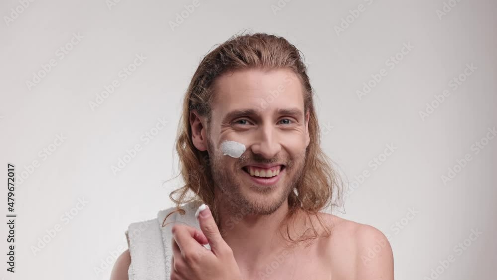 caucasian blond male with towel uses creams of his face and smiling. Handsome young curly man without shirt applying facial cream while look at the camera and smiling. Concept of healthcare and beauty caucasian blond male with towel uses creams of his face and smiling. Handsome young curly man without shirt applying facial cream while look at the camera and smiling. Concept of healthcare and beauty