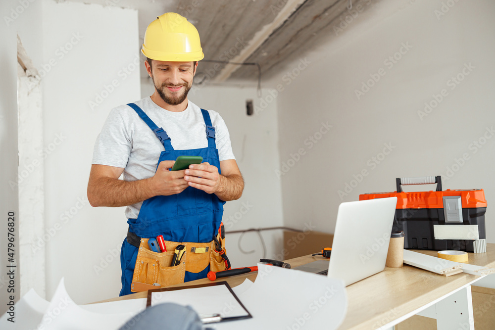 Joyful handyman in workwear and hardhat smiling while using smartphone, standing indoors during renovation work Joyful handyman in workwear and hardhat smiling while using smartphone, standing indoors during renovation work