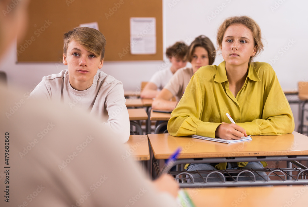 Obraz premium Teenage boy and girl listening to teacher and writing exercises in notebook at lesson in secondary school