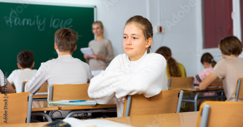 Sad teenage girl turned around while sitting at desk in classroom during lesson.