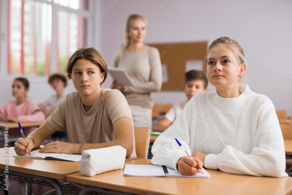 Teenage boy and girl sitting at table in classroom and working on tasks during lesson.