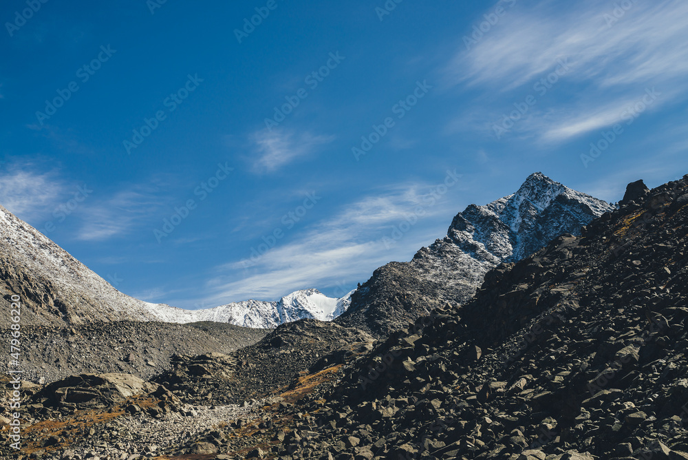 Awesome alpine landscape with dark rockies and high snow-covered mountains in sunshine under blue sky with cirrus clouds. Beautiful highland scenery with sharp black rocks and mountain top with snow.