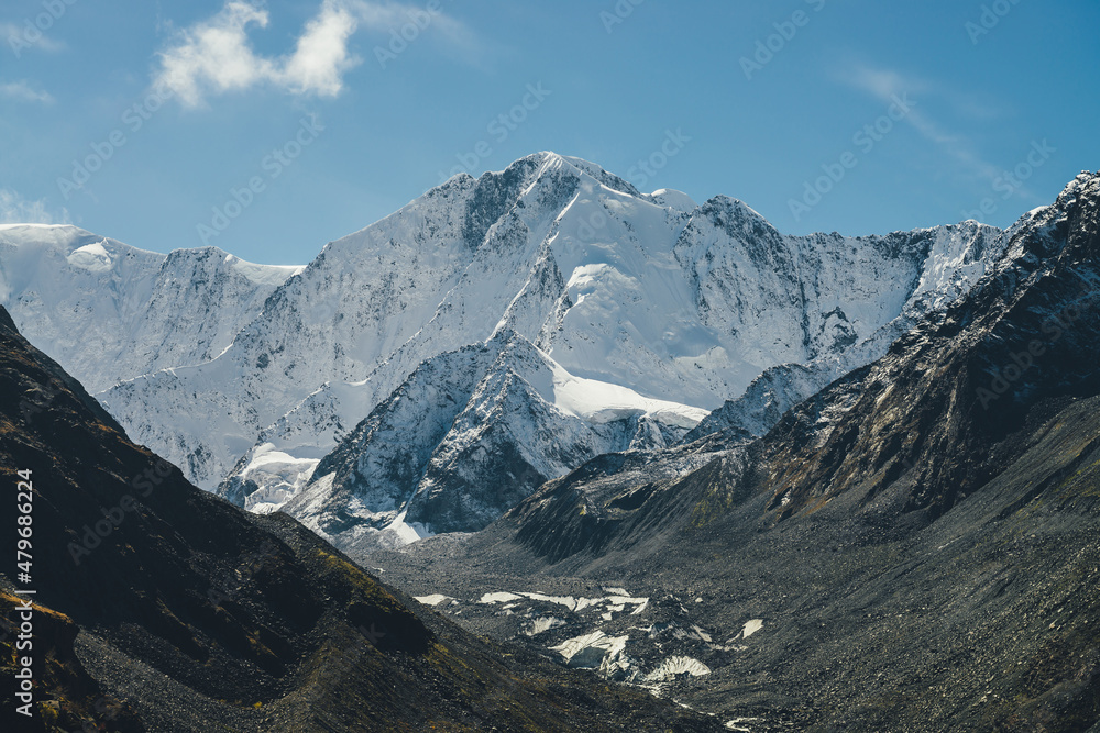 Alpine sunny landscape with high snowy mountain with peaked top and glacier between black rocks under cirrus clouds in sky. Big snow covered mountains in sunshine. White-snow pointy peak in sunlight.