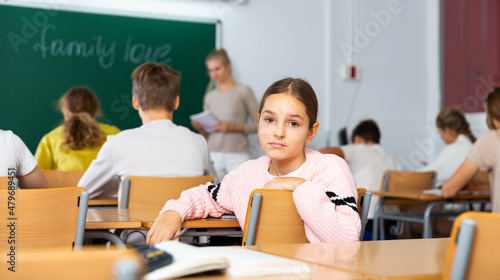 Sad young schoolgirl turned around while sitting at desk in classroom and looking at camera.