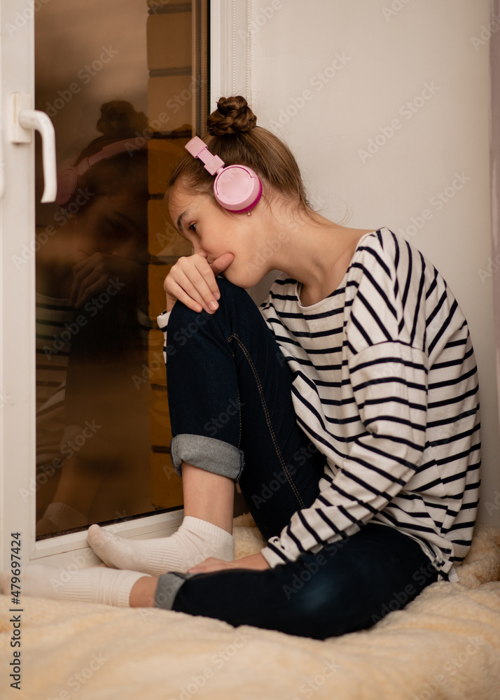 Fototapeta premium A cute teenage girl sits on a window listening to music with headphones and thinking.