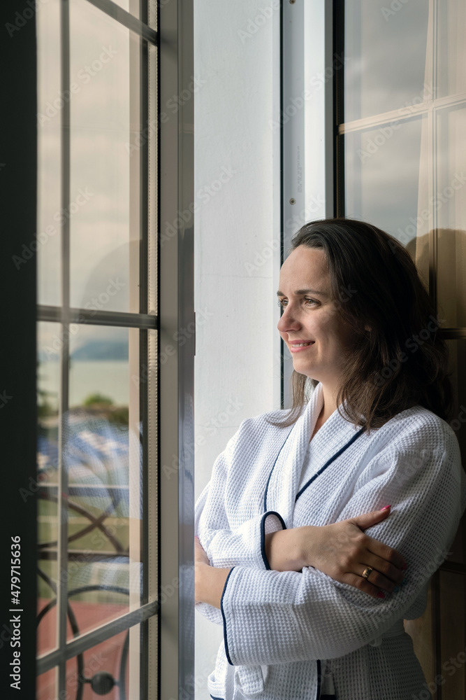 Smiling Caucasian Woman in White Bathrobe Enjoying the View while Standing in the Doorway of a Balcony of Hotel Room. Vacation and Travel Concept Smiling Caucasian Woman in White Bathrobe Enjoying the View while Standing in the Doorway of a Balcony of Hotel Room. Vacation and Travel Concept