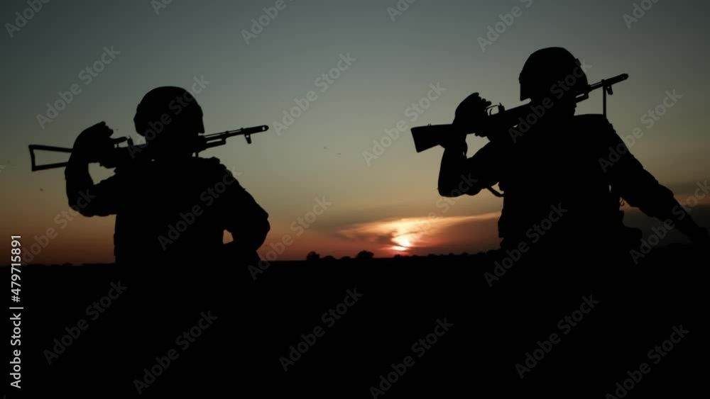Silhouettes of two soldiers with automatic rifle walking across field at sunset. Silhouettes of two soldiers with automatic rifle walking across field at sunset.