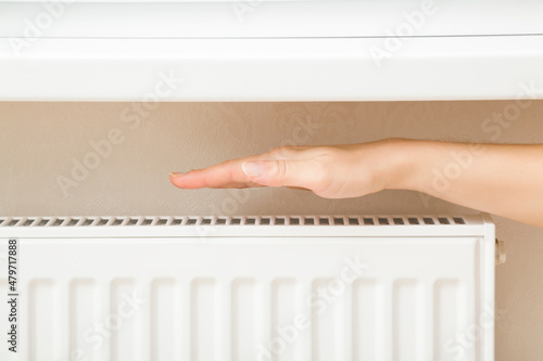 Young adult woman holding hand above white heater and checking warmth in room. Heating season. Closeup. Side view.