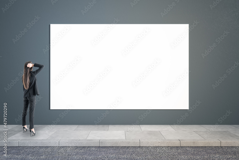 Thoughtful businesswoman looking at empty white poster in concrete interior. Mock up.