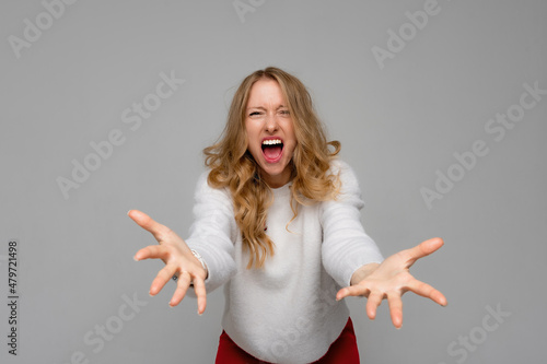 Irritated young woman screaming desperately with mouth wide open, looking at camera with angry expression, wears white sweater, standing over gray background