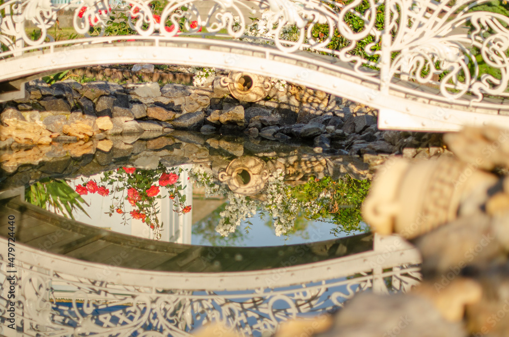 The decorative bridge and jug are reflected in the artificial pond. Beautiful summer photo of decorative garden decoration.