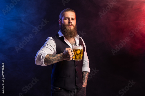 Half length of hipster bearded man who holds mug of beer looking at camera over smoke background