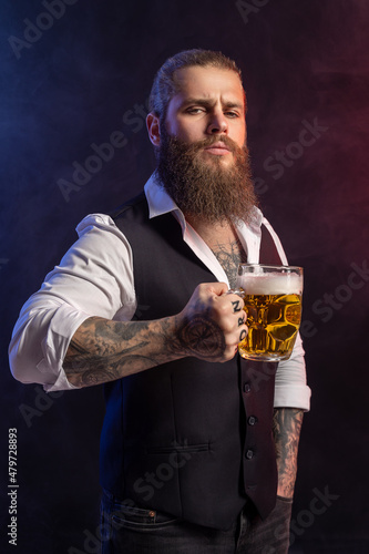 Portrait of Bearded man who holds mug of beer in his tattoos hand looking at camera over black background