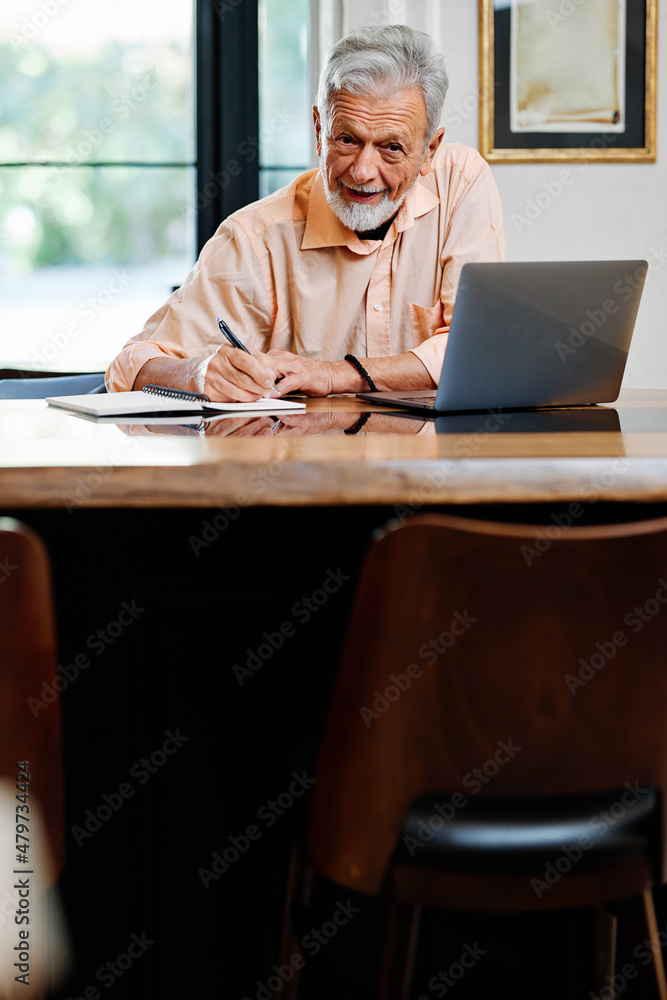 A happy senior student is sitting at home and writing down notes in a notebook while following an online lecture on the laptop. A happy senior student is sitting at home and writing down notes in a notebook while following an online lecture on the laptop.