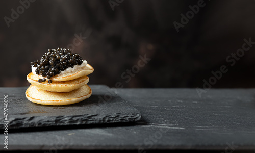A stack of blini pancakes with black caviar and sour cream on a slate, copy space photo
